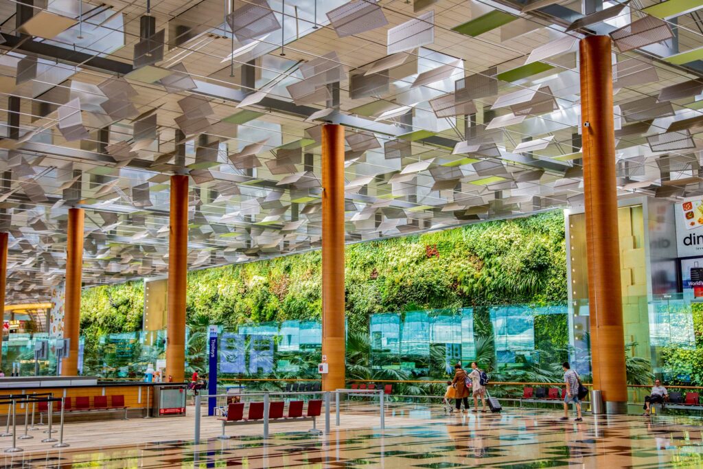 Changi Airport Terminal 3's interior boasts a stunning vertical garden, a lush tapestry of greenery adorning a towering glass wall. The garden is complemented by a soaring ceiling with artistic hanging panels and warm, wooden pillars, creating a harmonious blend of nature and modern architecture.