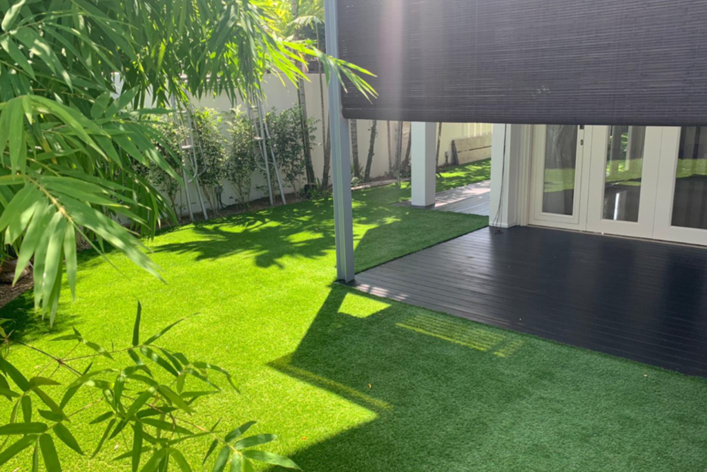 This backyard showcases a modern landscaping design featuring artificial turf, a dark wooden deck, and a black awning providing shade for glass doors. The keyboards are strategically placed to highlight the landscape company