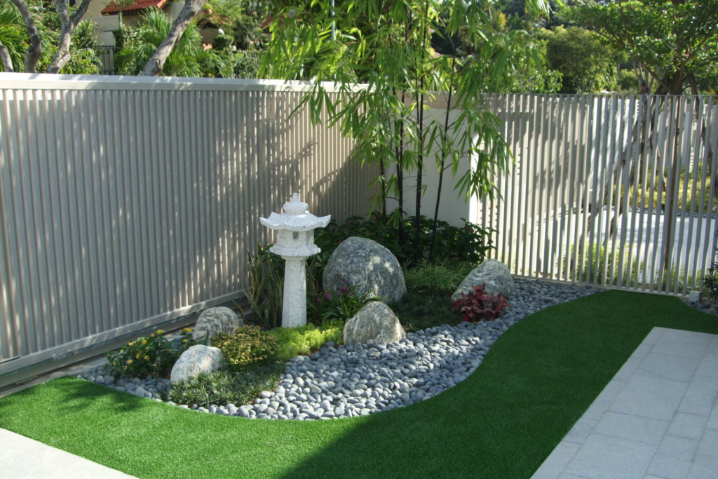 This meticulously designed garden features a Zen-like aesthetic with a stone lantern, bamboo, and carefully placed rocks. A bed of smooth pebbles adds texture, while the lush green artificial turf provides a low-maintenance contrast. A slatted fence provides privacy, completing the serene atmosphere.