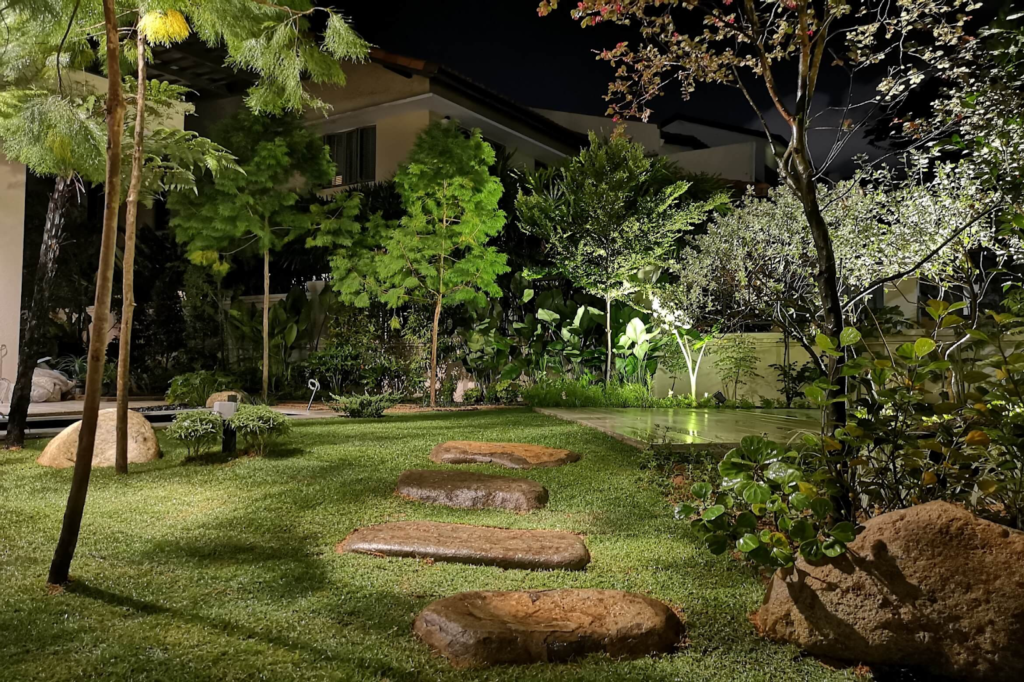 This meticulously landscaped garden at night features a lush green lawn, strategically placed spotlights illuminating a variety of trees and shrubs. A pathway of flat stones leads to a dimly lit patio, creating a serene and inviting atmosphere.