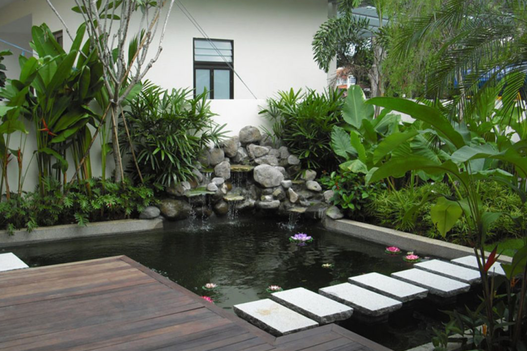 This backyard showcases a Hawaiian-inspired landscape design with a serene koi pond, a cascading rock waterfall, and lush tropical greenery. The wooden deck and stepping stones create a tranquil atmosphere, perfect for relaxation and enjoyment.